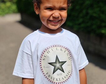 Puede incluir: Una camiseta blanca con un diseño de estrella roja, blanca y verde. La estrella tiene un contorno blanco y está rodeada de un círculo rojo y blanco con el texto "SCHULKIND" en letras negras. El texto "ERSTKASSIG" está debajo de la estrella y el año "2024" está en el centro de la estrella.