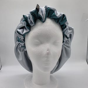 Pu&ograve; includere: Un cappuccio in raso grigio e verde acqua, progettato per proteggere i capelli. Il cappuccio presenta un bordo a balze ed &egrave; esposto su una testa di manichino bianca. Il raso gli conferisce un aspetto liscio e lussuoso.