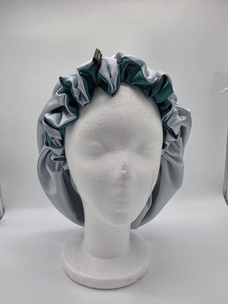 Pu&ograve; includere: Un cappuccio in raso grigio e verde acqua, progettato per proteggere i capelli. Il cappuccio presenta un bordo a balze ed &egrave; esposto su una testa di manichino bianca. Il raso gli conferisce un aspetto liscio e lussuoso.