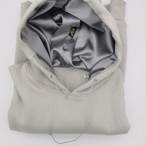 May include: A light gray hooded sweatshirt with a satin lining. The hood is folded down and the drawstring is visible. The sweatshirt has a small tag on the front with the letter "M" on it.