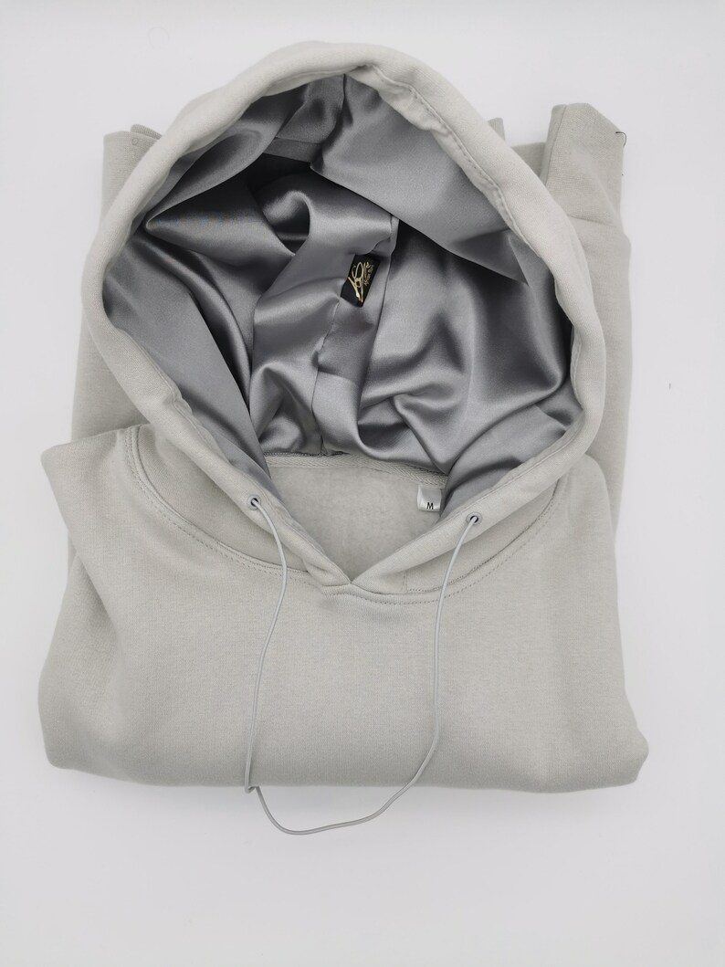 May include: A light gray hooded sweatshirt with a satin lining. The hood is folded down and the drawstring is visible. The sweatshirt has a small tag on the front with the letter "M" on it.