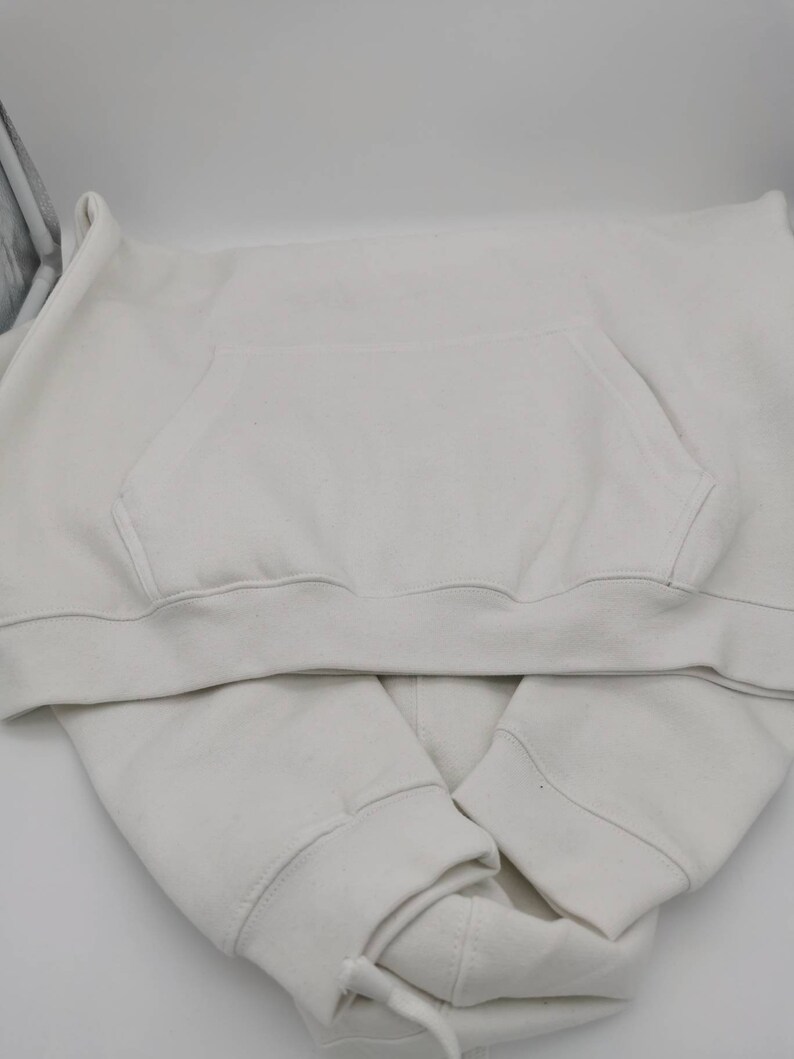 May include: A white hoodie with a pouch pocket on the front. The hoodie is folded in half and the hood is tucked inside.