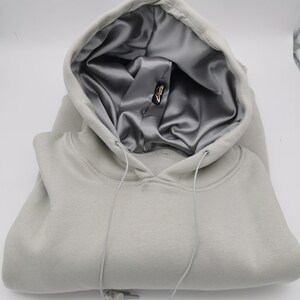May include: A gray hooded sweatshirt with a silver satin lining. The hood is folded down and the drawstring is visible. The sweatshirt is folded in half and the drawstring is hanging down.