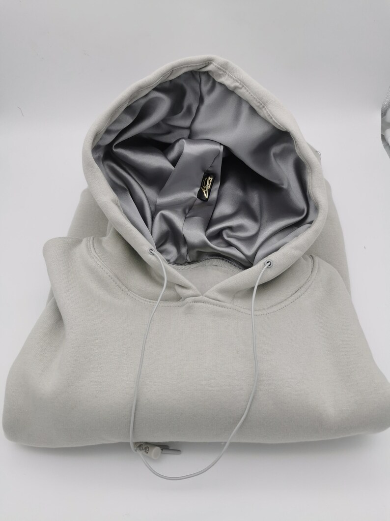 May include: A gray hooded sweatshirt with a silver satin lining. The hood is folded down and the drawstring is visible. The sweatshirt is folded in half and the drawstring is hanging down.