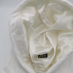 May include: White hooded sweatshirt with a white satin lining. The hood is pulled up and the lining is visible. A small black tag with the text "African Storm" is visible on the lining.