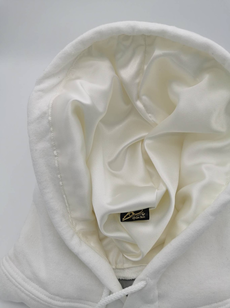 May include: White hooded sweatshirt with a white satin lining. The hood is pulled up and the lining is visible. A small black tag with the text "African Storm" is visible on the lining.