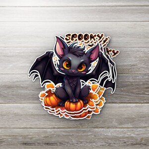 May include: A black bat with orange wings sits on top of a pile of orange pumpkins. The bat has large, round eyes and a cute, cartoonish expression. The text "Spooky" is written in black letters above the bat.