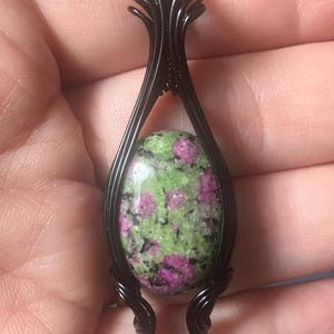 May include: A pendant featuring an oval-shaped ruby zoisite gemstone with mottled green and magenta-pink coloring, encased in a decorative black wire-wrapped metal frame that extends upward into a bail for hanging on a chain or cord.