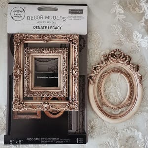 May include: A package of Decor Moulds with the text "Ornate Legacy" and a decorative frame. The package is 5" x 8". Next to the package is a decorative oval frame. The frames are a light brown color.
