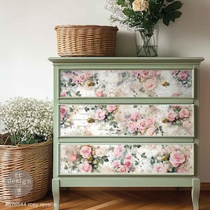 May include: A sage green dresser with three drawers, each adorned with a floral rose design. The roses are pink and white, set against a vintage-style background. Gold knobs and a woven basket on top complete the look. The text "#676544 rosy reverie" is at the bottom.