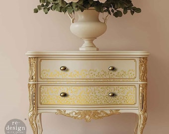 May include: A white two-drawer dresser with gold accents and ornate detailing. The drawers have a gold leaf pattern and are accented with brass knobs. The dresser is sitting on a light pink surface.