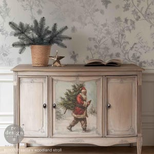 May include: A light-colored wooden cabinet with three doors, featuring a painted image of Santa Claus carrying a Christmas tree. The cabinet is decorated with a star and a basket with a plant on top. The text on the bottom says "santa's woodland stroll".