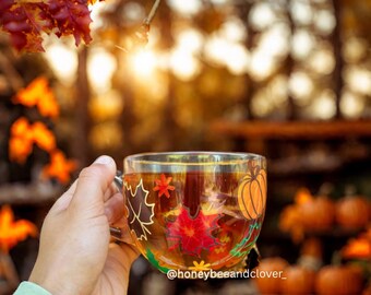May include: A clear glass mug filled with tea, decorated with fall-themed leaf and pumpkin designs. The mug is held up against a blurred background of autumn foliage and pumpkins. The text "@honeybeeandclover_" is visible on the mug.
