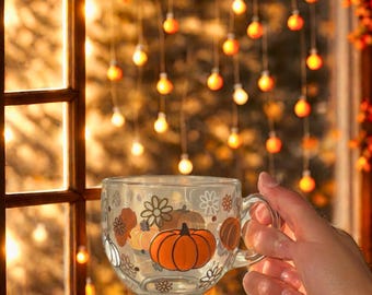 May include: A clear glass mug with a pumpkin and floral design. The mug is filled with a warm beverage and held in front of a window with string lights. The mug has a handle and is decorated with orange pumpkins and white flowers.