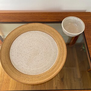 Könnte beinhalten: Ein großer, gesprenkelter Keramikservierteller mit breitem, strukturiertem Rand, kombiniert mit einer kleineren Schüssel. Der Teller hat eine hellbeige Farbe mit einem gesprenkelten weißen Zentrum. Die Schüssel ist ebenfalls gesprenkelt.