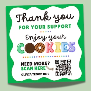 May include: A square green and white sign with the words "Thank you for your support" and "Enjoy your cookies." The word "cookies" is in colorful letters. The sign also includes a QR code and the text "Need more? Scan here."