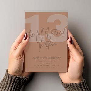Puede incluir: Una tarjeta de invitación rectangular con un fondo marrón claro es sostenida por dos manos. La tarjeta presenta el número 13 en un color marrón claro, con las palabras "the fall I turned thirteen" en una fuente de escritura. Debajo está el texto para el 13 cumpleaños de Isabel.