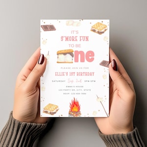May include: A birthday invitation with a s'mores theme. The invitation features the text "It's S'more Fun to be One" in pink, along with illustrations of s'mores, marshmallows, and a campfire. The invitation is held by a person.