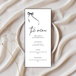 Puede incluir: Una tarjeta de menú blanca con texto negro y una ilustración de lazo en la parte superior. El menú dice "The Menu" y enumera un entrante, un plato principal y un postre para el 30 cumpleaños de Megan. La tarjeta está sobre un plato blanco.