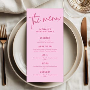 May include: A pink menu card with the words "the menu" in cursive script. The menu is for Megan's 30th birthday and lists the starter, appetizer, main, sides, and dessert. The menu is on a white plate with gold silverware.