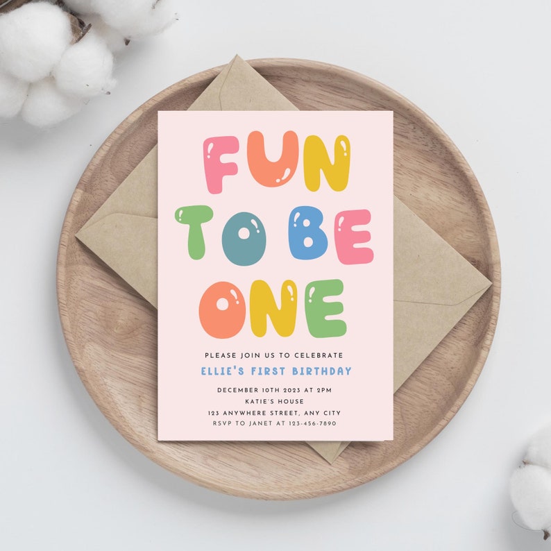 Może przedstawiać: R&oacute;żowe zaproszenie na przyjęcie urodzinowe z tekstem "FUN TO BE ONE" w kolorowych literach w kształcie baniek. Zaproszenie zawiera szczeg&oacute;ły dotyczące pierwszych urodzin Ellie, w tym datę, godzinę, miejsce i informacje RSVP.