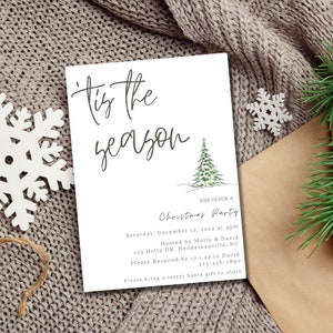 May include: A white card with the text 'Tis the season' in black cursive script. The card is decorated with a watercolor illustration of a green pine tree. The text below the illustration reads 'Join us for a Christmas Party' followed by details about the party, including the date, time, host, address, and RSVP information. The card is lying on a brown knitted blanket with a white snowflake ornament.