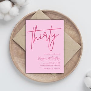 May include: A pink birthday invitation with the word "thirty" in a cursive font. The invitation includes details for Maggie's 30th birthday celebration, including the date, time, and location. The invitation is placed on a wooden tray with a brown envelope.