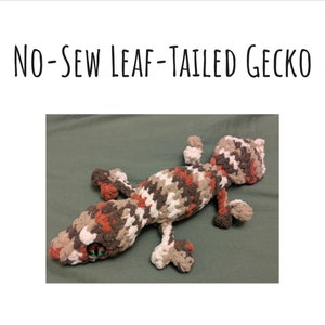May include: A crocheted stuffed gecko with a brown, white and orange pattern. The gecko has a large, green eye and a long, curled tail.