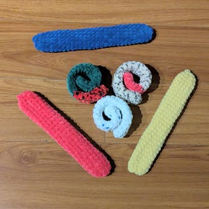Può includere: Una collezione di articoli all'uncinetto fatti a mano in varie forme e colori. Include un lungo rettangolo blu, un rettangolo rosso, un rettangolo giallo e tre pezzi a spirale in verde, bianco e azzurro. Gli articoli sono disposti su una superficie di legno.