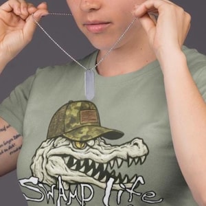 Può includere: Una collana con targhetta d'argento viene tenuta davanti a una persona che indossa una maglietta verde. La maglietta presenta una grafica di un alligatore con un cappello mimetico, con le parole "SWAMP LIFE" in bianco.