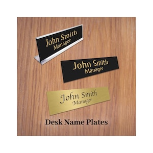 May include: Three desk name plates displaying "John Smith, Manager". One is black with a silver stand, another is black, and the third is gold. The text "Desk Name Plates" is at the bottom, all on a wooden surface.