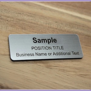 May include: A rectangular silver-toned metal name badge with rounded corners. The badge features black printed text that reads Sample, POSITION TITLE, and Business Name or Additional Text, arranged in three distinct lines on the surface.
