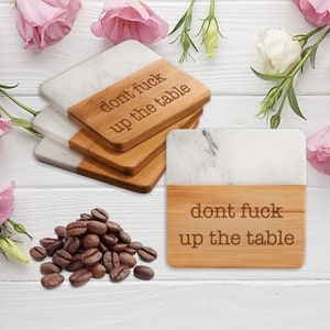 May include: Set of four coasters with a white marble top and a bamboo base. The coasters have the text "don't fuck up the table" engraved on the bamboo base.