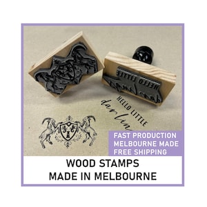May include: Two rectangular wooden stamps with black rubber bases. One stamp features an intricate crest design with two goats flanking a shield. The other stamp displays the text HELLO LITTLE DARLIN in a script and sans-serif font combination.