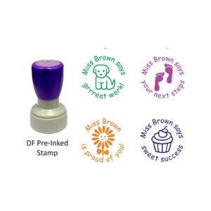 May include: A DF pre-inked stamp with a purple handle and grey base. Four circular stamp impressions are shown: a green puppy with 'Miss Brown says grrreat work!', purple footprints with 'Miss Brown says your next steps', an orange flower with 'Miss Brown is proud of you!', and a blue cupcake with 'Miss Brown says sweet success'.