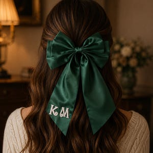 Può includere: Un fiocco per capelli in raso verde scuro con due code lunghe, caratterizzato dalle lettere KM ricamate in bianco sulla coda sinistra. Il fiocco è fissato tra i lunghi capelli castani ondulati e il tessuto ha una finitura liscia e lucida.