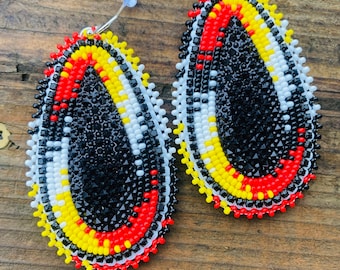 May include: A pair of oval-shaped beaded earrings with a black, white, red, and yellow color scheme. The earrings are made with small, round beads and have a traditional Native American design.