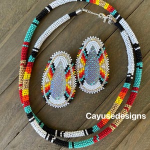 May include: A white beaded necklace with turquoise, red, black and gold accents. The necklace is paired with matching earrings with a white, turquoise, red, black and gold beaded design. The earrings have a silver iridescent centre.