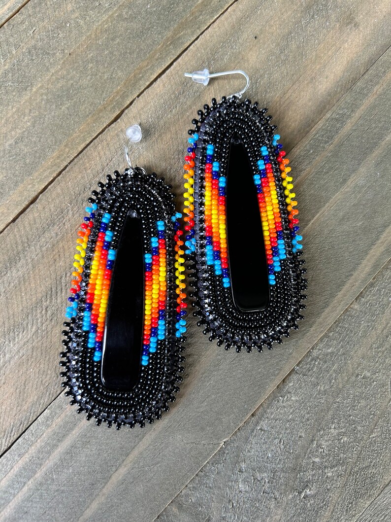 May include: A pair of black beaded earrings with colorful geometric patterns. The earrings are oval shaped and have a black backing.