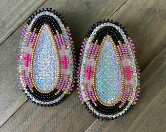 May include: A pair of iridescent beaded earrings with a teardrop shape. The earrings are black, white, pink, and gold with a sparkly center.