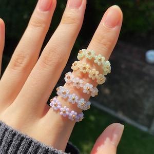 Puede incluir: Anillos de flores de cuentas en varios colores pastel: verde claro, amarillo, blanco, azul y morado. Cada anillo presenta pequeñas cuentas doradas en el centro del diseño floral. Los anillos se muestran en un dedo.
