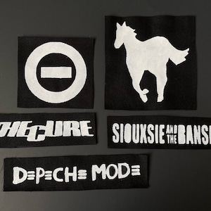 May include: A collection of black and white patches featuring band logos and symbols. The patches include the logos for The Cure, Siouxsie and the Banshees, Depeche Mode, and a white horse silhouette. The patches are rectangular and square.