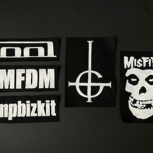 May include: A collection of black and white patches featuring band logos and symbols. The patches include logos for Tool, KMFDM, Limp Bizkit, a cross symbol, and the Misfits skull logo. The patches are rectangular and made of fabric.