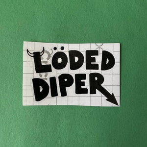 May include: A black and white sticker with the words "LÖDED DIPER" in a bold, playful font. The design includes a small devil horn graphic above the "Ö" and an arrow pointing down from the word "DIPER". The sticker is on a grid background against a green backdrop.