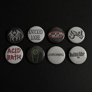 May include: A collection of eight round pinback buttons with various band logos and text. The buttons feature a range of colors, including black, white, and red. The text on the buttons includes band names such as "Knocked Loose" and "Acid Bath".