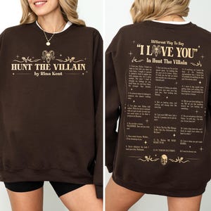 May include: A brown sweatshirt with the text "HUNT THE VILLAIN" and "by Rina Kent" in a gold font on the front. The back of the sweatshirt has the text "Different Way To Say 'I LOVE YOU' In Hunt The Villain".