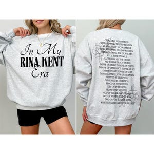 May include: Light gray sweatshirt with the text "In My Rina Kent Era" on the front. The back of the sweatshirt has a list of phrases and words, including "Cruel King" and "Black Knight".