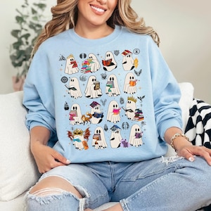 Pode incluir: Uma camisola de gola redonda azul-clara com uma impressão gráfica repetida de vários fantasmas brancos, cada um interagindo com vários livros, pequenos animais e itens mágicos, dispostos num padrão de grelha na frente da peça.