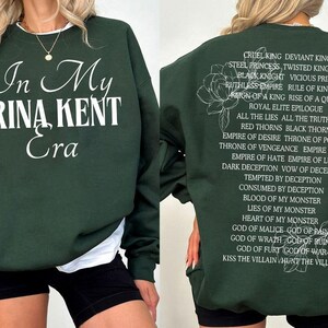 May include: Dark green sweatshirt with the text "In My Rina Kent Era" in white script on the front. The back features a list of phrases and a floral design in white. A casual, comfortable style.