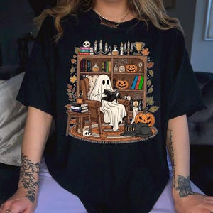 Könnte beinhalten: Ein schwarzes T-Shirt mit einer grafischen Illustration eines weißen Geistes, der in einem braunen Sessel sitzt und ein Buch liest. Die Szene zeigt ein hölzernes Bücherregal mit Büchern, Kürbissen, Kerzen, Totenköpfen und einer schwarzen Katze.